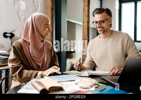Two multinational colleagues designers smiling while working with laptop computer at workshop indoors Stock Photo