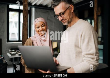 Two multinational colleagues smiling while working with laptop computer in modern office Stock Photo
