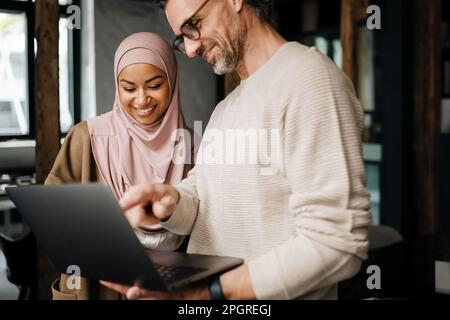 Two multinational colleagues smiling while working with laptop computer in modern office Stock Photo