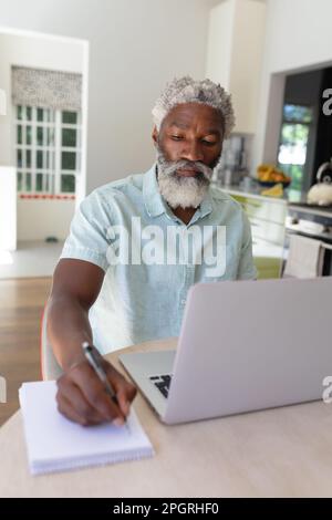 African american senor man making notes and using laptop in kitchen. staying at home in isolation during quarantine lockdown. Stock Photo