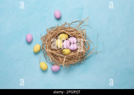 Banner for the Easter holiday. Colored eggs in a straw nest and white spring flowers on a blue background. Place for text. Top view. Stock Photo