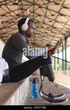 Fit, mixed race disabled male athlete at an outdoor sports stadium, sitting in the stands wearing headphones using smartphone wearing running blades. Stock Photo