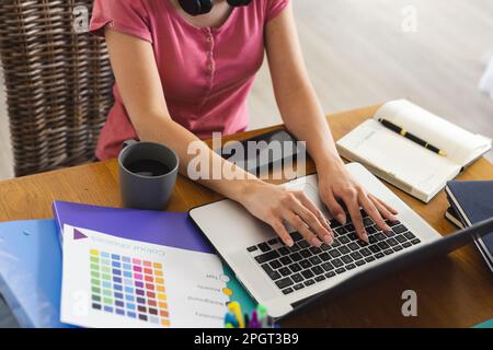 Midsection of caucasian young woman with color swatch and files working over laptop on table. Unaltered, hand, coffee, small business, home office, te Stock Photo