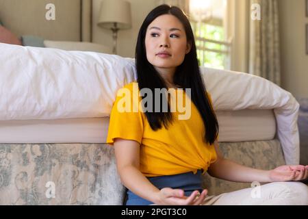 Thoughtful asian woman sitting on floor meditating in bedroom, looking away. Healthy lifestyle, free time and relaxation. Stock Photo