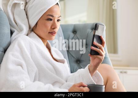Happy asian woman wearing bathrobe and towel on head sitting in armchair using smartphone. Lifestyle, self care, communication, health and beauty. Stock Photo