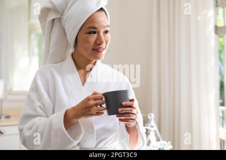 Happy asian woman wearing bathrobe and towel on head drinking coffee in bathroom. Lifestyle, self care, relaxation, health and beauty. Stock Photo