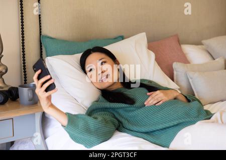 Smiling asian woman lying on bed with head on pillows using smartphone. Lifestyle, free time, communication and relaxation. Stock Photo