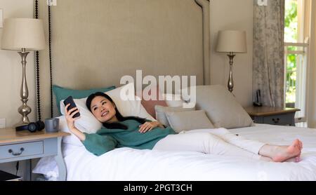 Smiling asian woman lying on bed with head on pillows using smartphone. Lifestyle, free time, communication and relaxation. Stock Photo