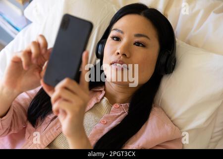 Happy asian woman lying on bed wearing headphones using smartphone. Lifestyle, free time, communication and relaxation. Stock Photo