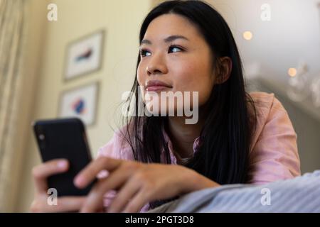 Thoughtful asian woman lying on bed using smartphone, looking away and smiling. Lifestyle, free time, communication and relaxation. Stock Photo