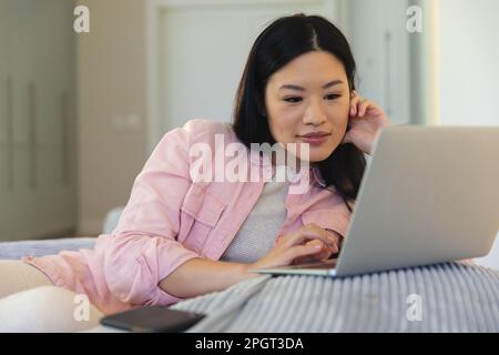 Happy asian woman sitting on beanbag at home using laptop, smiling. Lifestyle, relaxation and communication. Stock Photo