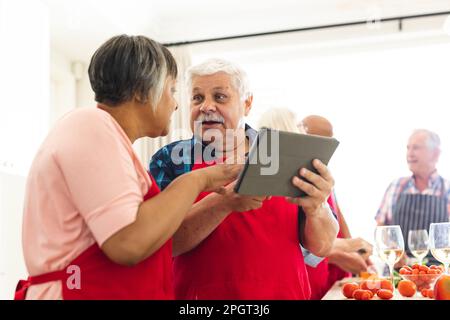 Happy group of diverse senior friends in aprons, using tablet during cookery class. Retirement, learning, cooking and healthy lifestyle. Stock Photo