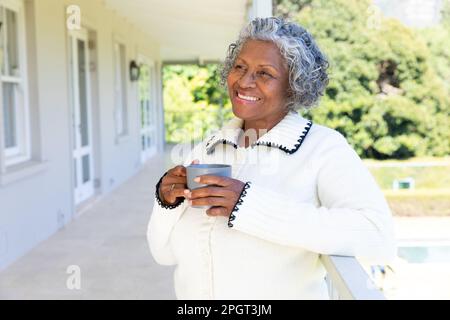 Thoughtful smiling african american senior woman with coffee cup standing by railing in balcony. Copy space, unaltered, lifestyle, drink, looking away Stock Photo