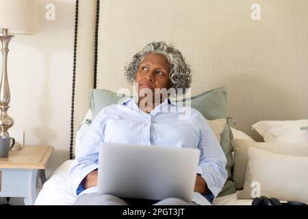African american senior businesswoman contemplating while working over laptop on bed, copy space. Unaltered, professional, work from home, pensive and Stock Photo