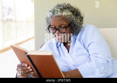 Happy african american senior woman reading book while relaxing on sofa at home. Copy space, unaltered, lifestyle, eyeglasses, smile, knowledge, hobby Stock Photo