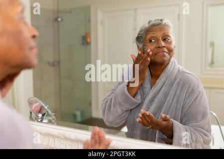 African american senior woman dressed in bathrobe looking in mirror and applying face cream at home. Unaltered, body care, aging, reflection, pamperin Stock Photo