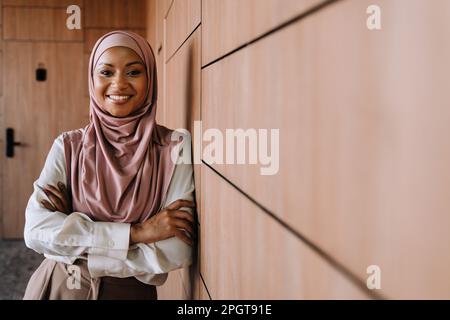 Happy muslim businesswoman smiling and standing by wall while working in office Stock Photo