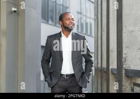 Portrait of a successful senior African-American businessman, lawyer, banker standing near a business center in a suit, holding his hands in his pockets, looking to the side with a smile. Stock Photo