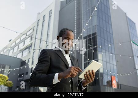 A senior African American businessman is talking to his team via video call. Standing near an office center in a suit, wearing headphones and holding a tablet. Stock Photo