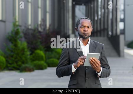 Portrait of a successful black senior businessman, investor, lawyer standing near an office center, holding a tablet in his hands. He looks seriously into the camera. Stock Photo