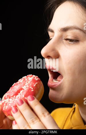 A woman bites a large red donut, a black background, a place for text. Gluttony, overeating and sugar addict. Stock Photo