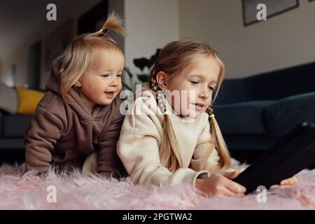 Caucasian siblings using tablet at home. Distance or remote learning for child. Pretty stylish schoolgirl with her baby sister watching cartoons Stock Photo