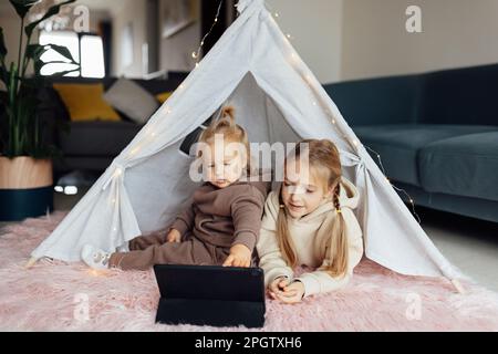 Caucasian siblings using tablet at home. Distance or remote learning for child. Pretty stylish schoolgirl with her baby sister watching cartoons Stock Photo