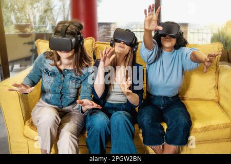Three young women, two Caucasian and one Brazilian, aged 20-30, sitting on a couch in a gaming lounge, wearing VR headsets and using hand tracking tec Stock Photo