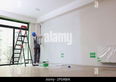 Employee with helmet paints wall with roller in premise Stock Photo
