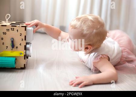 Cute little girl is playing with busiboard outdoors on green grass. Educational toy for toddlers. girl opened door to cube of board. Stock Photo