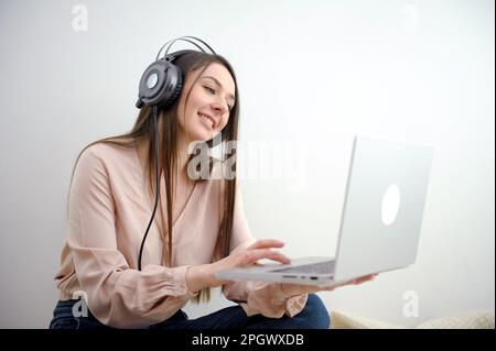Cheery Caucasian woman in headphones having online video call on laptop, sitting on chair Millennial lady participating in webinar, communicating remotely on internet nice smile camera slowly zooms Stock Photo