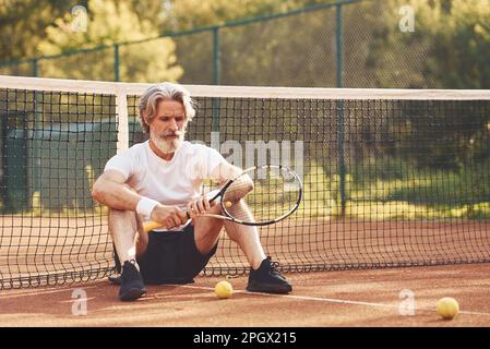 Sitting and taking a break. Senior stylish man in white shirt and black sportive shorts on tennis court Stock Photo