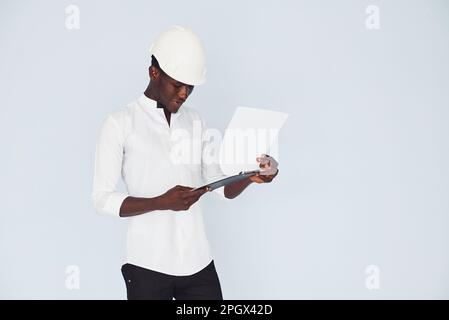 Young african american engineer in white formal clothes standing against wall indoors with documents Stock Photo