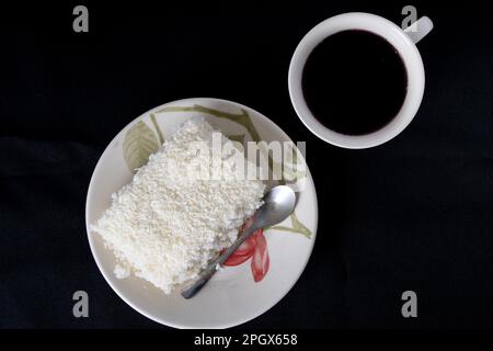 Piece of couscous in porcelain saucer on black background, typical Brazilian dish, made with tapioca, milk, sugar and coconut milk. With cup of coffee Stock Photo