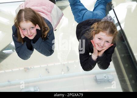 Tourist children child kid visitors to Tower Bridge pose to camera for photographst whilst lying on glass floors floor window in the high level walkways / walk way that joins the two towers. River Thames and car deck can be seen below. London, UK. (133) Stock Photo