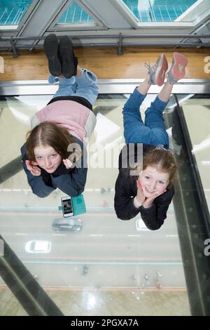 Tourist children child kid visitors to Tower Bridge pose to camera for photographst whilst lying on glass floors floor window in the high level walkways / walk way that joins the two towers. River Thames and car deck can be seen below. London, UK. (133) Stock Photo