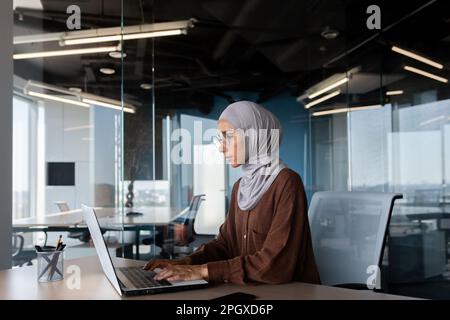 Serious and thinking woman in hijab working inside office with laptop, Muslim female programmer typing on keyboard writing software code at workplace working artificial intelligence development. Stock Photo