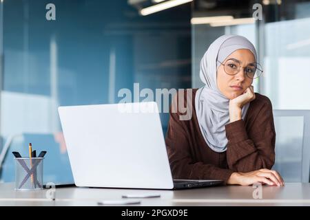 Sad disappointed woman in hijab looking at camera, businesswoman at workplace not satisfied with achievement results, upset muslim woman working inside office with laptop. Stock Photo