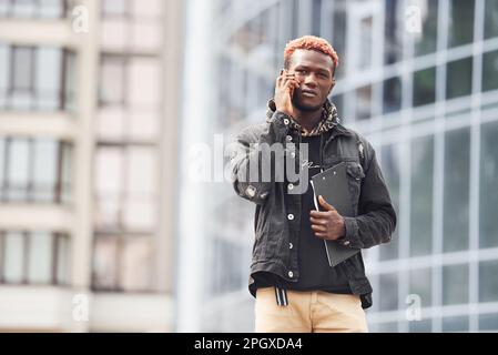 Talking by phone. Young african american man in black jacket outdoors in the city standing against modern business building Stock Photo