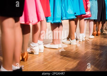 Young girls ballroom dancers standing in a row before beginners competition. Colorful dresses and different shoes on wooden floor. Stock Photo