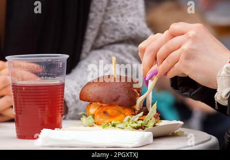 Young lady enjoying a fresh burger at the farmers street food market, close-up of hands holding single-use cutlery and burger with drink Stock Photo