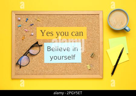Corkboard with motivational quotes, cup of coffee and office stationery on yellow background, flat lay Stock Photo