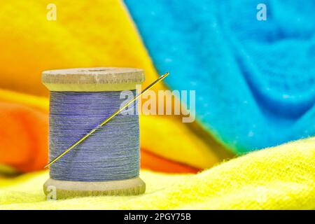Spool of purple cotton thread and sewing needle sitting on bright colored clothing. Textile tailoring concept with focus on foreground. Stock Photo