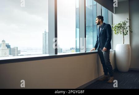 My business dreams are constantly manifesting. a young businessman looking thoughtfully out of an office window. Stock Photo