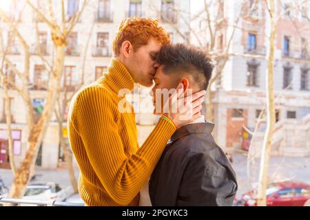 Beautiful gay couple being romantic at home on the sofa, home balcony kissing each other on the forehead, lgbt concept Stock Photo