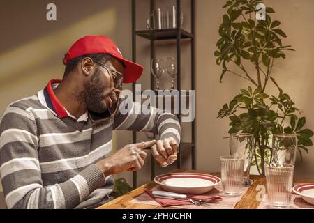 Young African man in casual clothes and red backwards hat, sitting at a restaurant table set for two with an empty chair, talking on the phone and poi Stock Photo