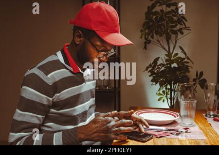 Young African man in casual clothes and red cap sitting at a restaurant table browsing content on his smartphone, laid on the table - people technolog Stock Photo