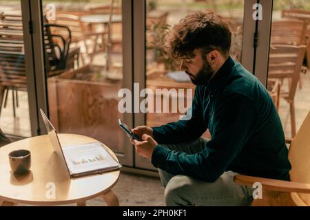 Handsome well-dressed young man scrolling through social media on his phone and working on the laptop at the cafe Stock Photo