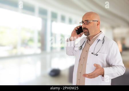 Serious male doctor talking on cell phone, standing in hospital. Physician in white uniform with stethoscope. Confident therapist. Stock Photo