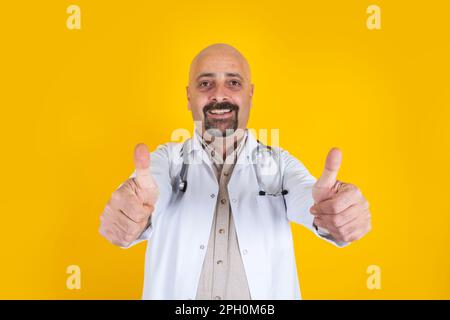 caucasian middle aged doctor happy thumbs up. Isolated yellow studio background. Looking camera, approving, successful treatment concept idea. Stock Photo